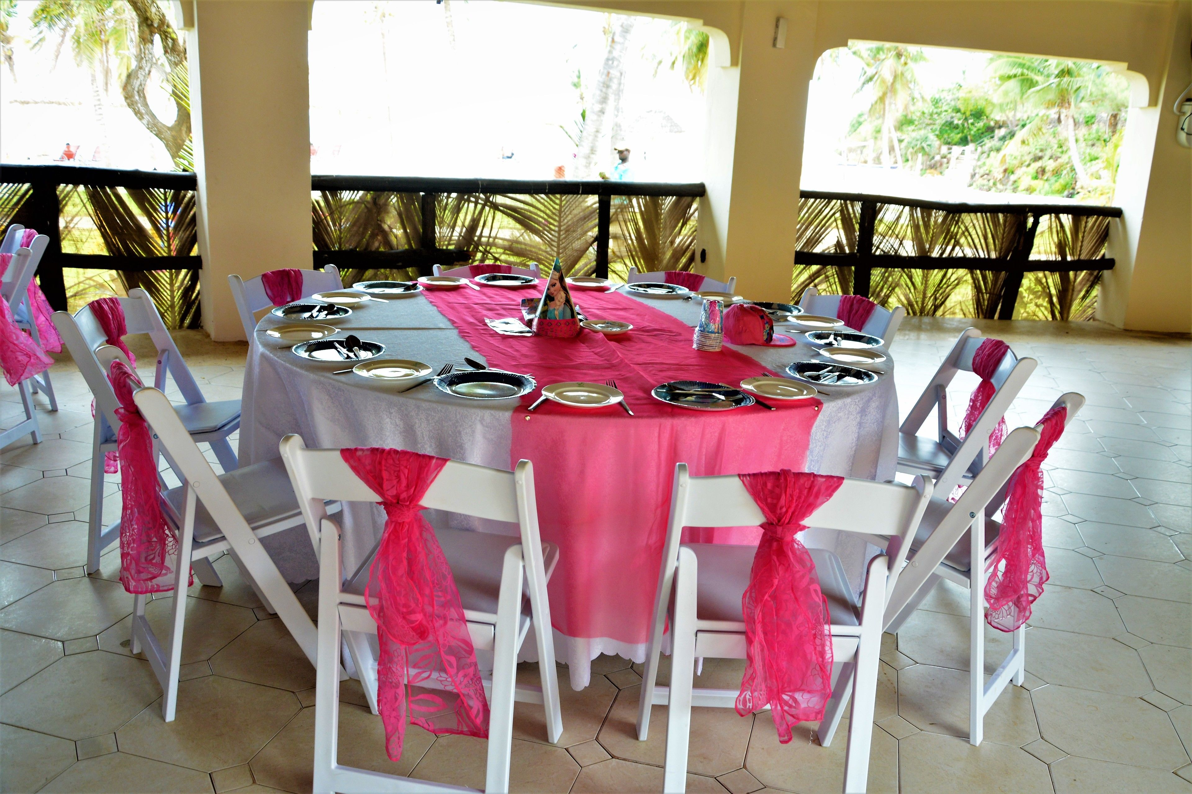 Mesa redonda dispuesta para una comida, con sillas blancas, mantel rosa y vistas al exterior con vegetación.