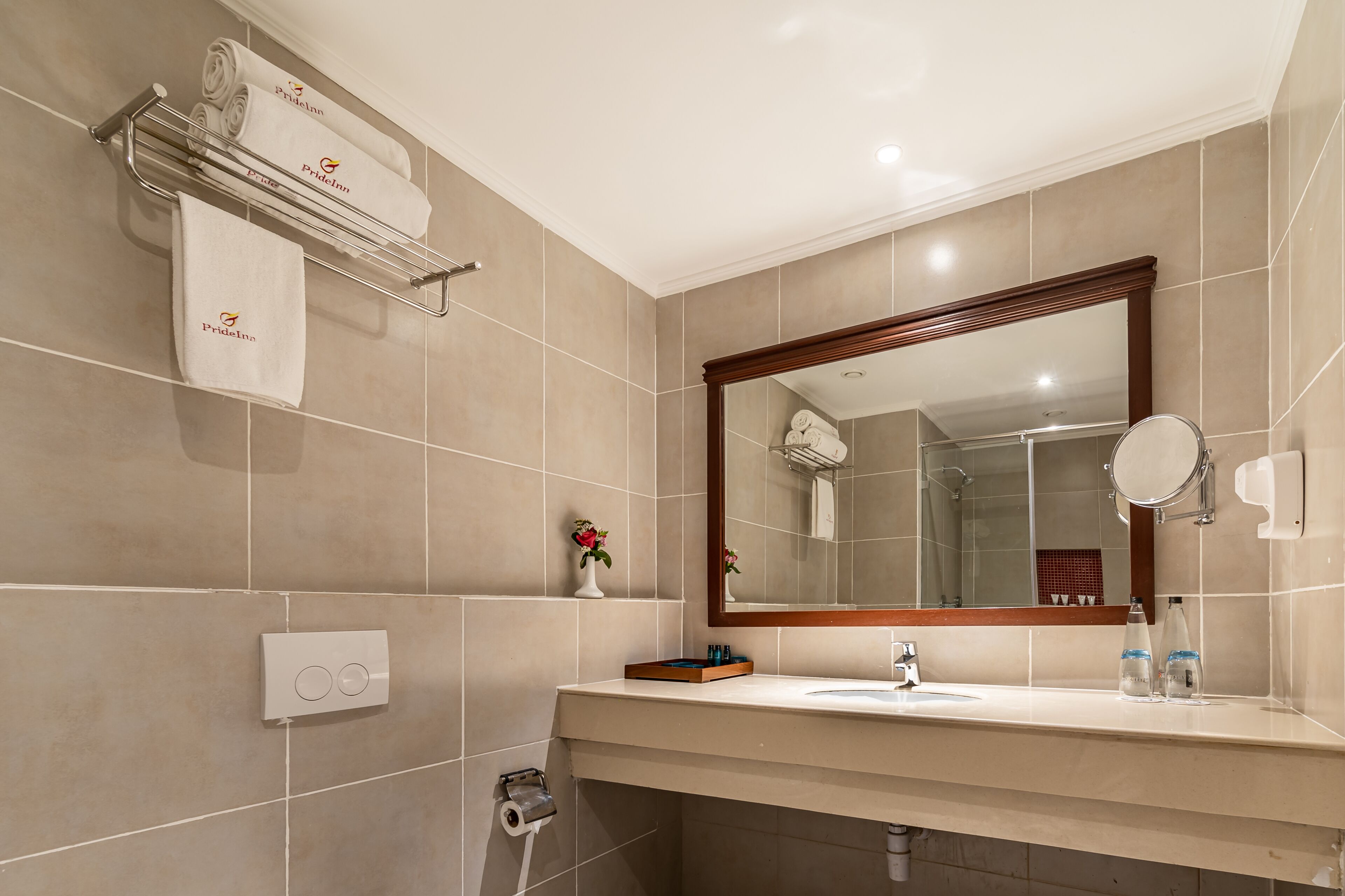 Un baño moderno con azulejos color beige, un espejo grande, un perchero con toallas de la marca, un lavamanos con artículos de aseo y un dispensador de papel higiénico.