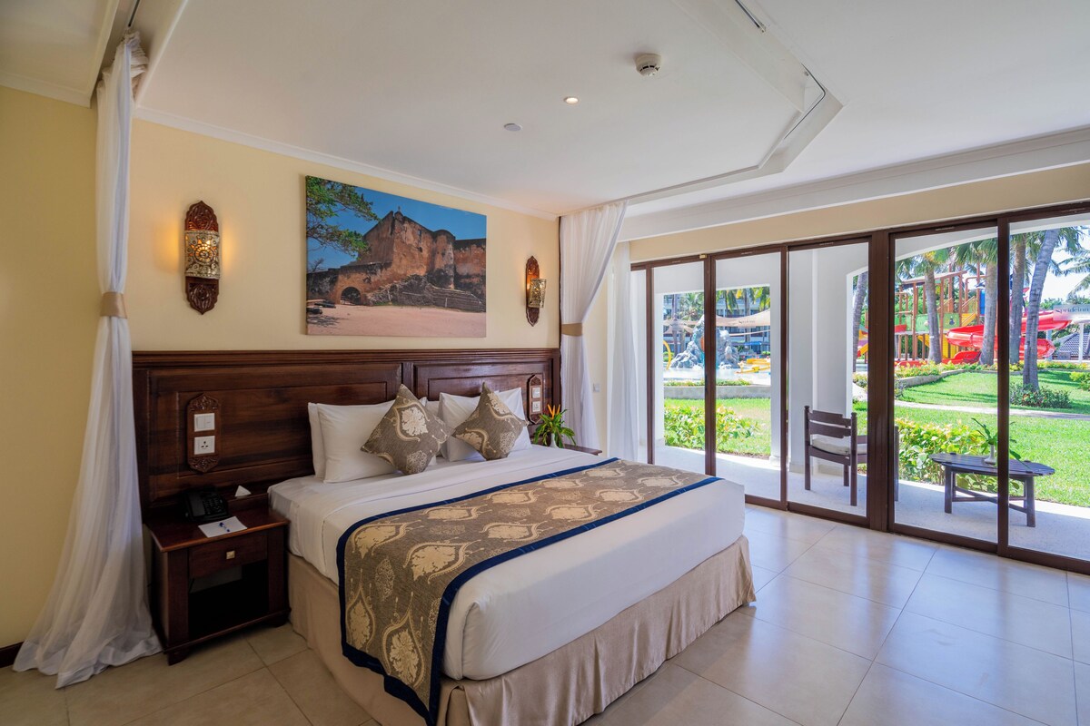 Habitación de hotel con una cama grande, una mesita de noche, un cuadro en la pared y vistas a un parque acuático a través de la puerta de cristal.