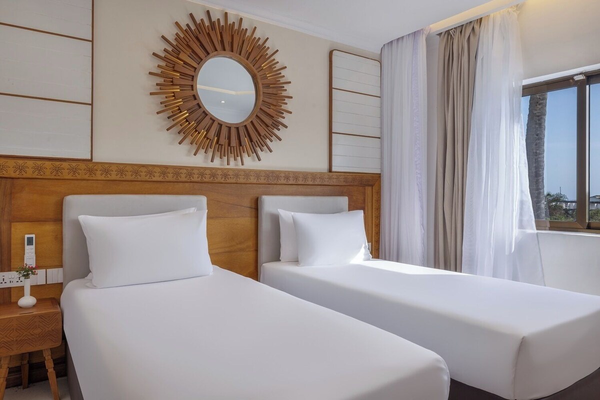Habitación de hotel con dos camas, un espejo grande, una mesita de noche y una ventana con cortinas.