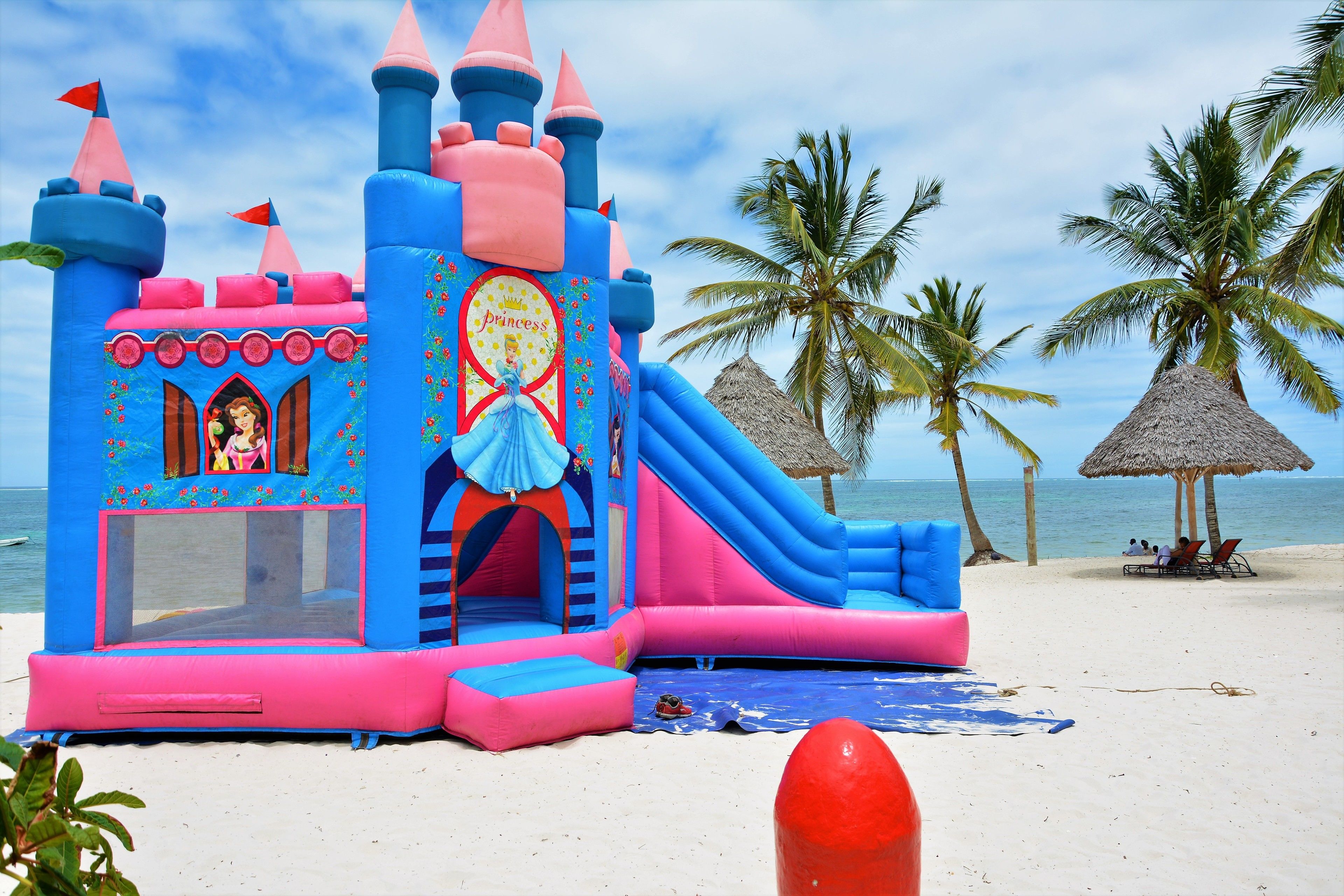 Un castello gonfiabile a tema principessa sulla spiaggia, con uno scivolo e palme sullo sfondo.