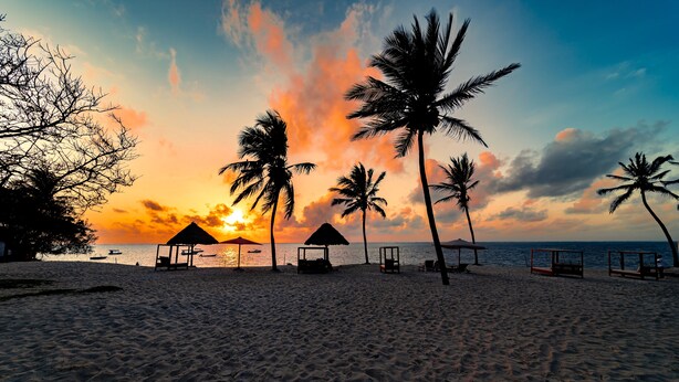 Uma praia com palmeiras, cabanas de praia e o oceano ao pôr do sol.