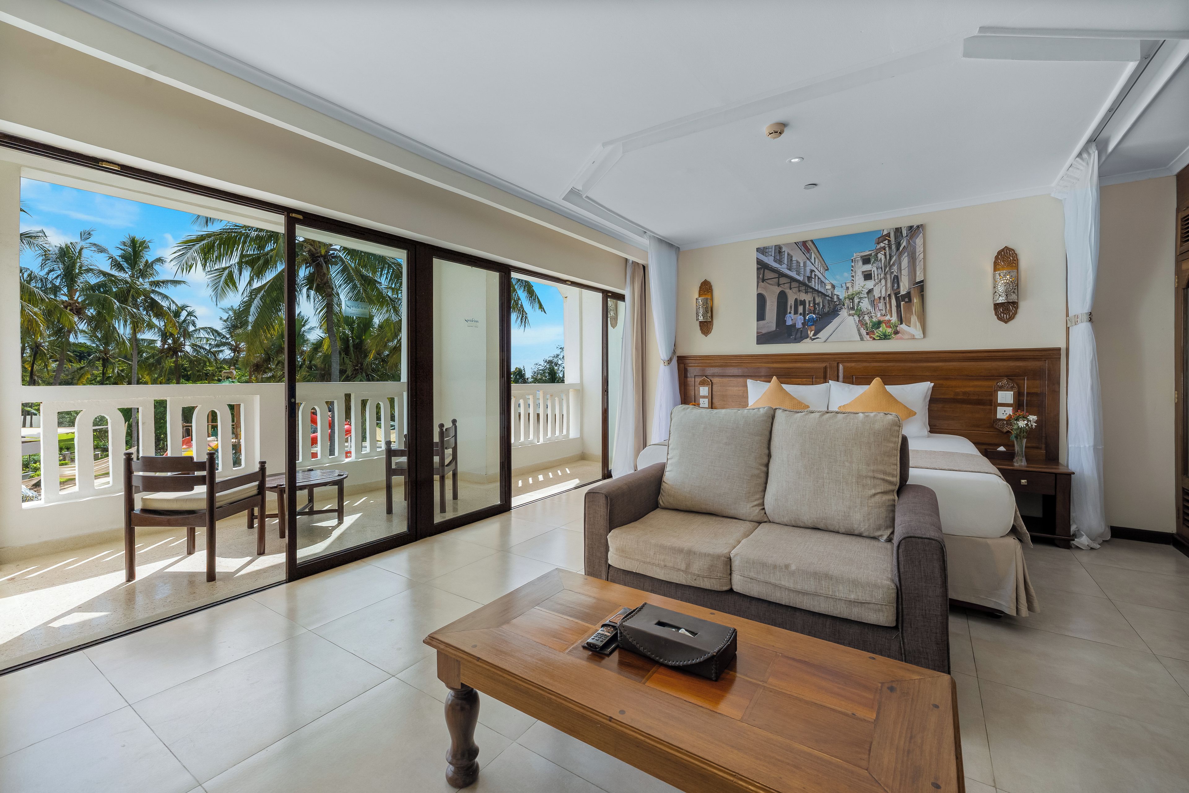 A hotel room with a balcony, a bed with pillows, a sofa, a coffee table, and a painting on the wall.
