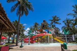 Un parque acuático con toboganes y zona de piscina, rodeado de palmeras y un edificio con techo de paja.