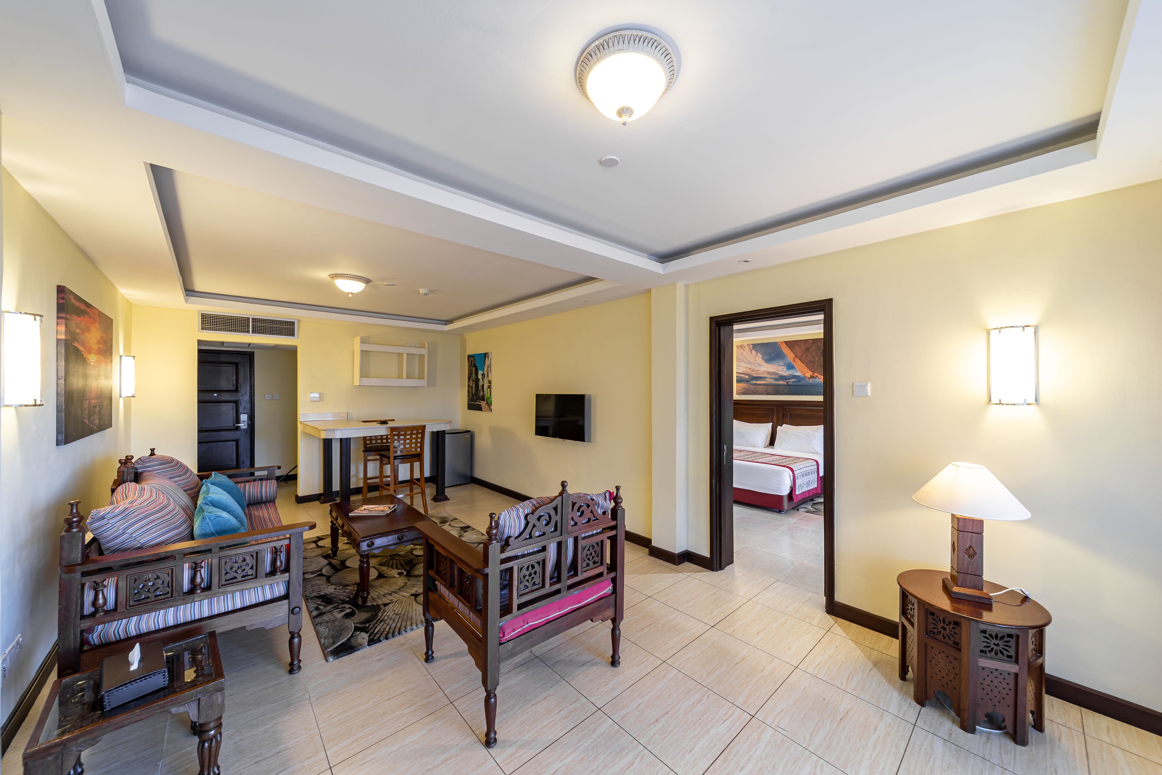 Un grand salon comprenant un canapé, une table basse et une lampe. Un téléviseur est fixé au mur et une porte donne accès à une autre pièce.