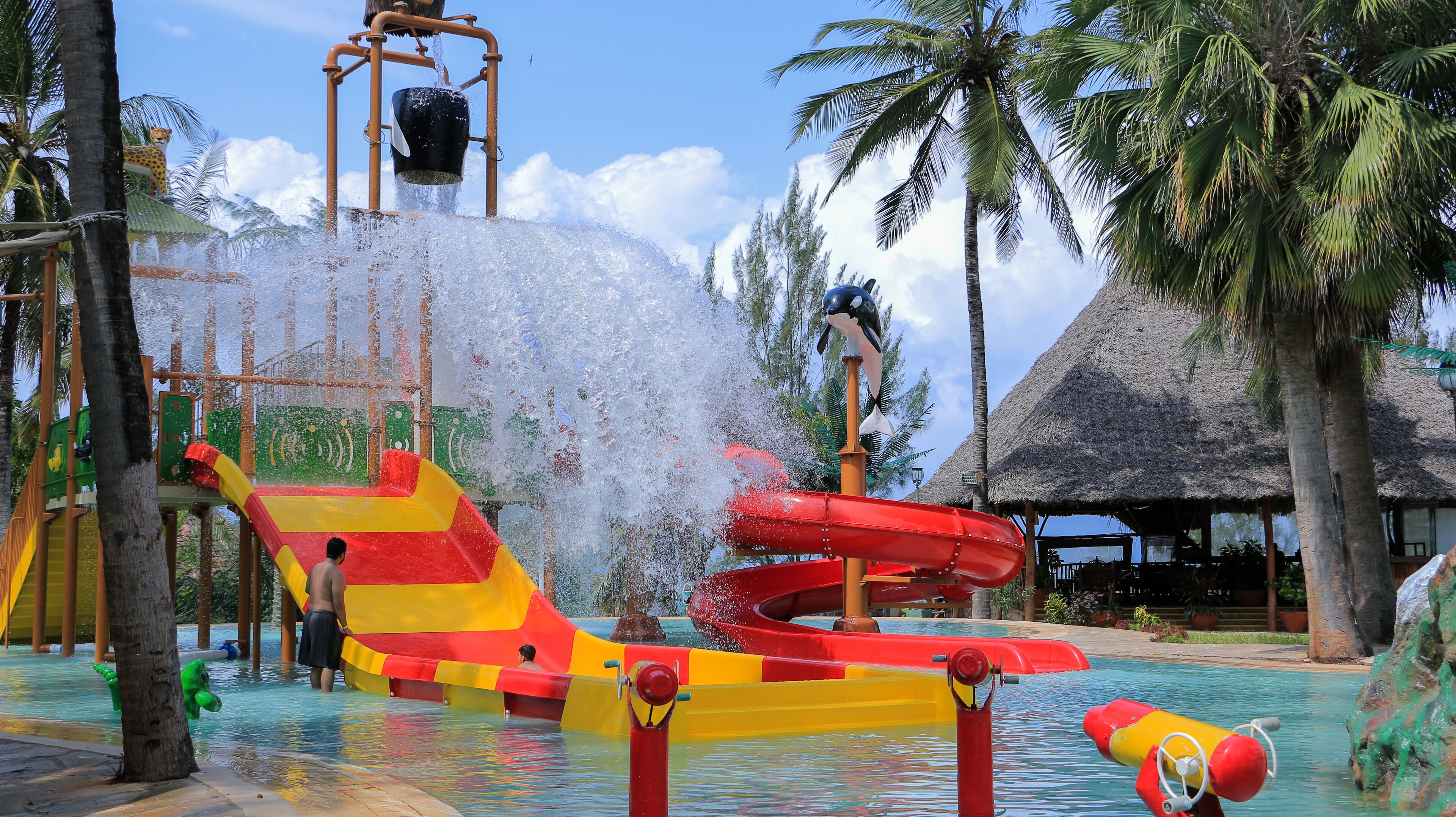 A water park with slides and water features, surrounded by palm trees and a thatched-roof structure.