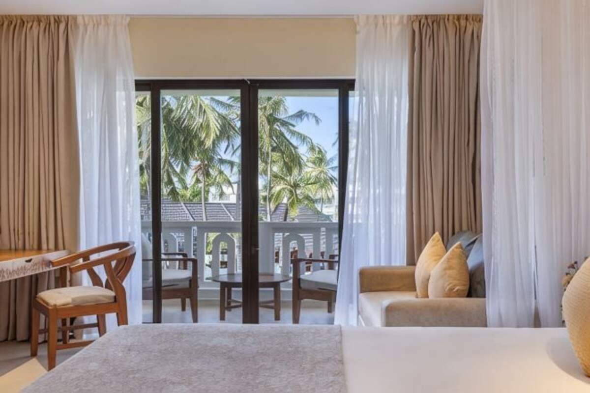 A hotel room with a bed, a seating area with chairs, a table, and a balcony with palm trees.