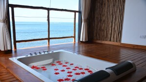 Uma banheira grande com flores vermelhas flutuando na água, piso de madeira e vista para o mar através de uma janela.