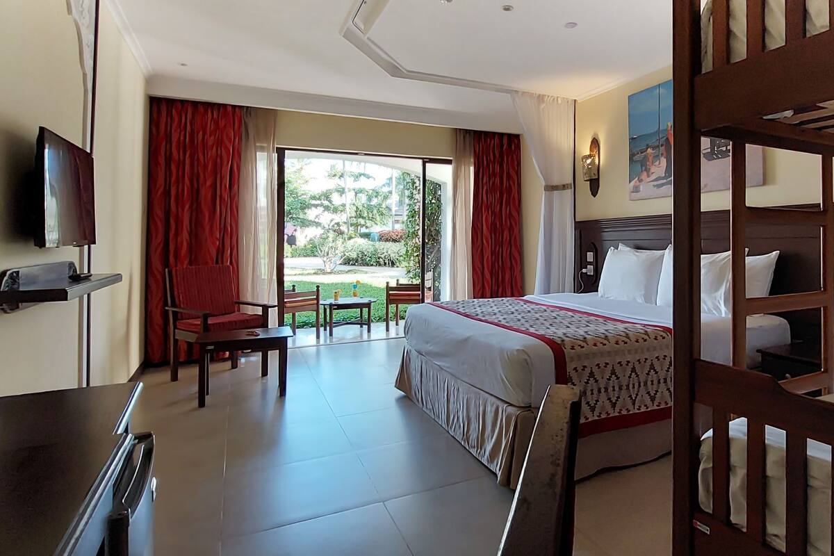 Habitación de hotel con una cama grande, una cama más pequeña, televisión, escritorio y balcón con cortinas rojas.