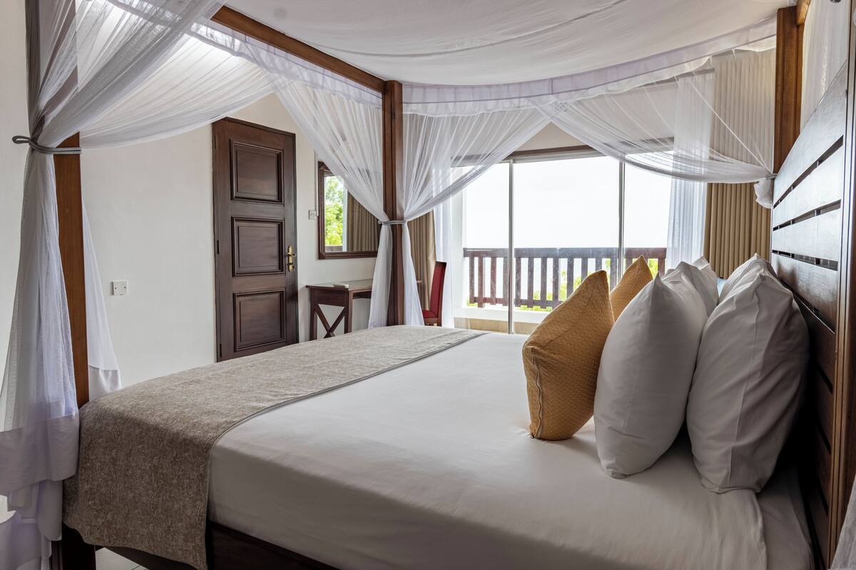 Un dormitorio con cama, ropa de cama blanca, cabecera de madera, mesita de noche, escritorio y ventana con vistas al exterior.