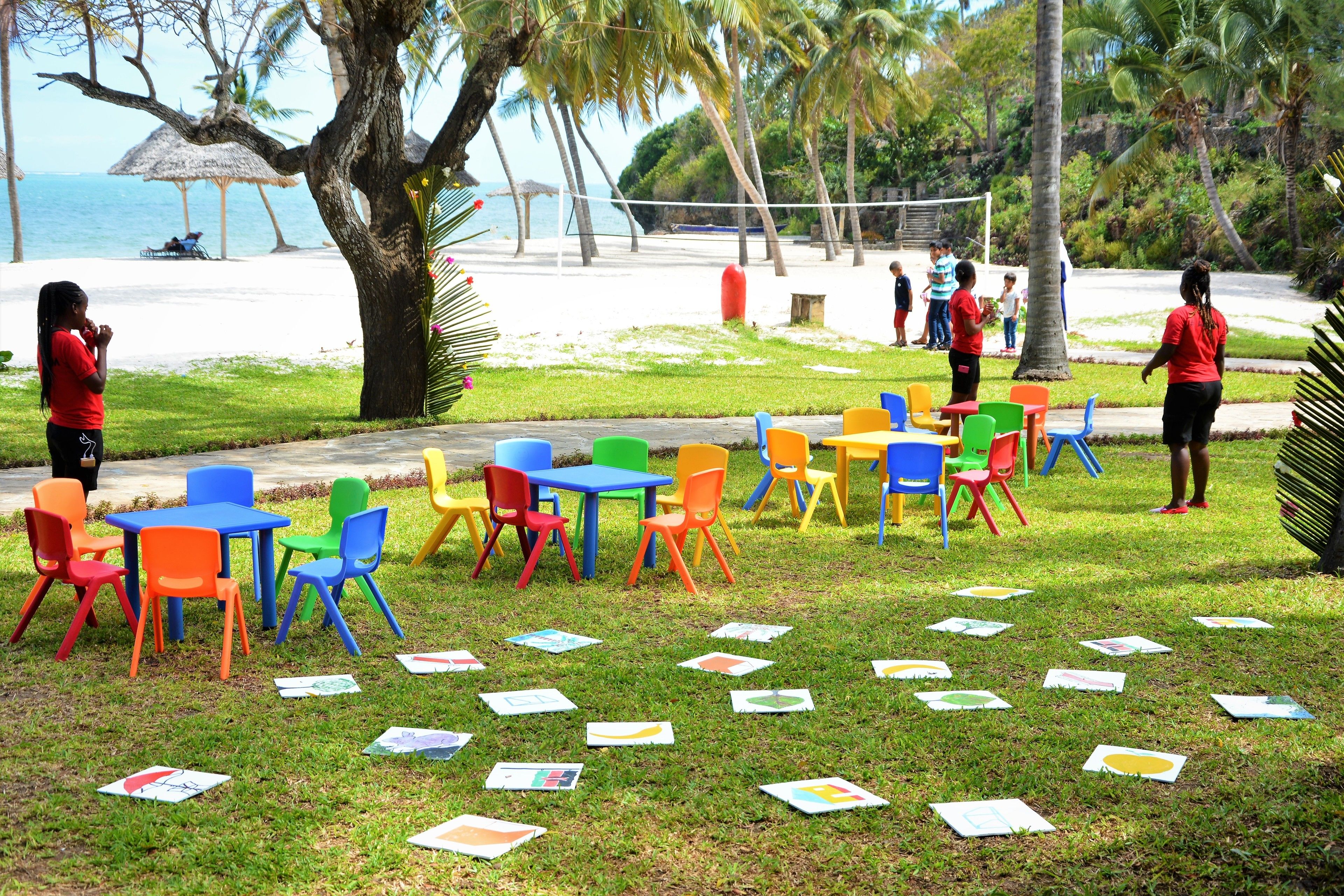 A grassy area with colorful plastic chairs and tables, scattered papers, and a beach in the background.