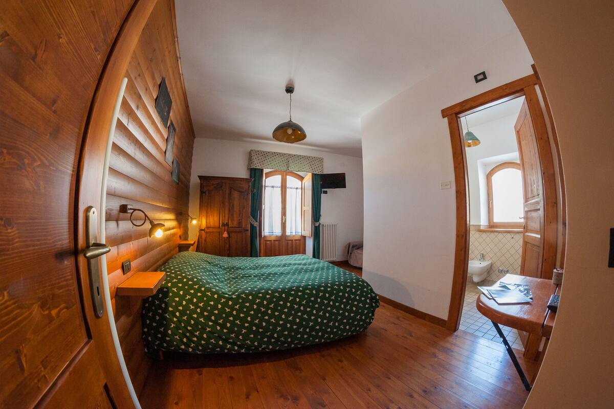 Une chambre à coucher avec une tête de lit en bois, un lit recouvert d’une couverture à pois verts, une table de chevet en bois, un bureau avec une chaise et une fenêtre avec des rideaux.