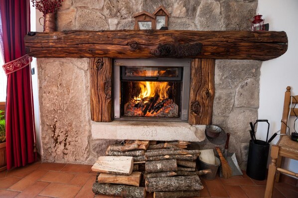 A stone fireplace with a wooden mantel, a fire burning inside, and a stack of firewood in front.