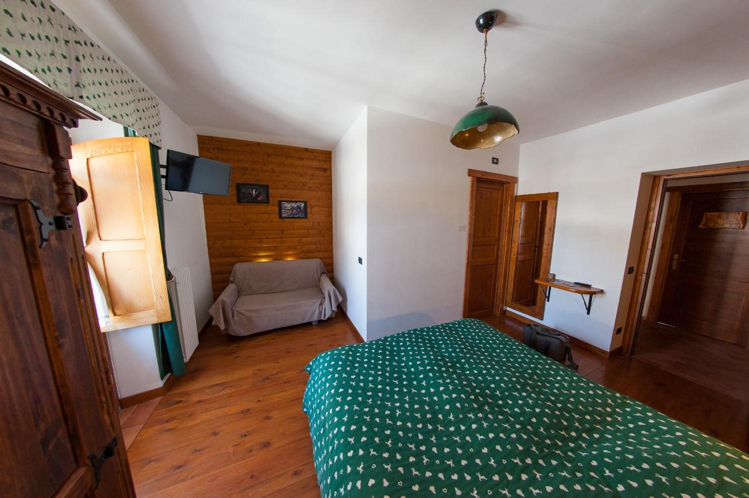 A room with wooden flooring, a green patterned bed, a wooden wardrobe, a brown sofa, a mounted TV, and a green pendant light.