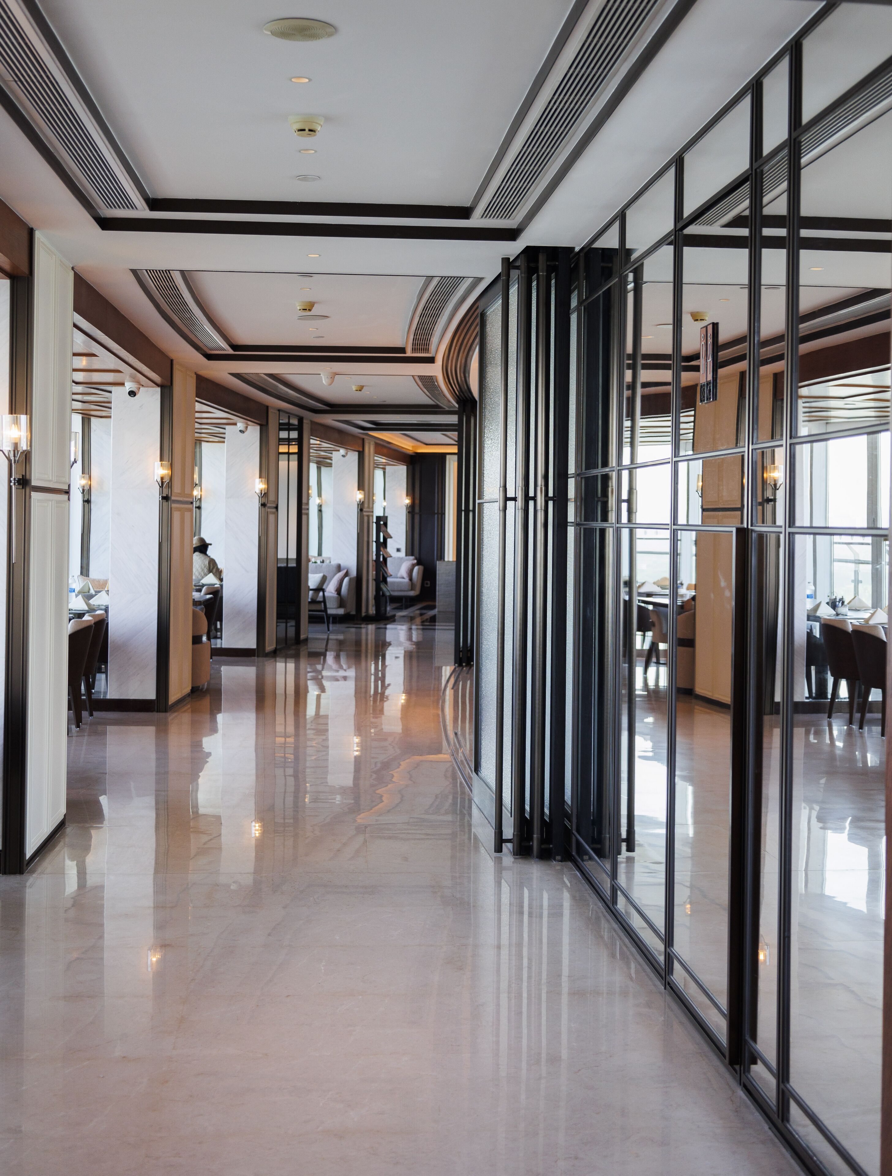 A modern hotel corridor with reflective floors, large windows, and seating areas.
