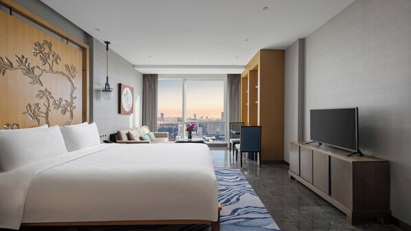 A modern hotel room with a large bed, a flat-screen TV, a seating area with a view of the city, and a large window.