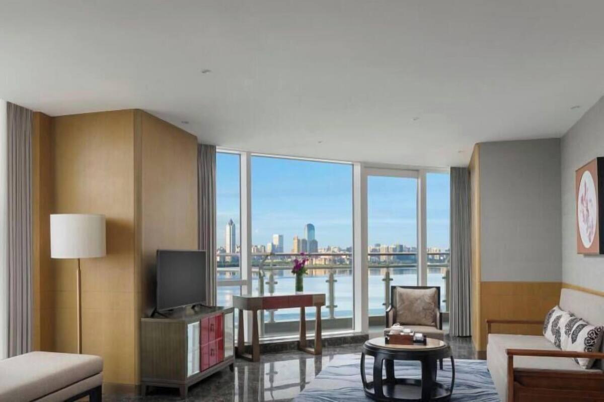 A modern hotel room with a large window, a flat-screen TV, a sofa, a coffee table, and a view of a city skyline.