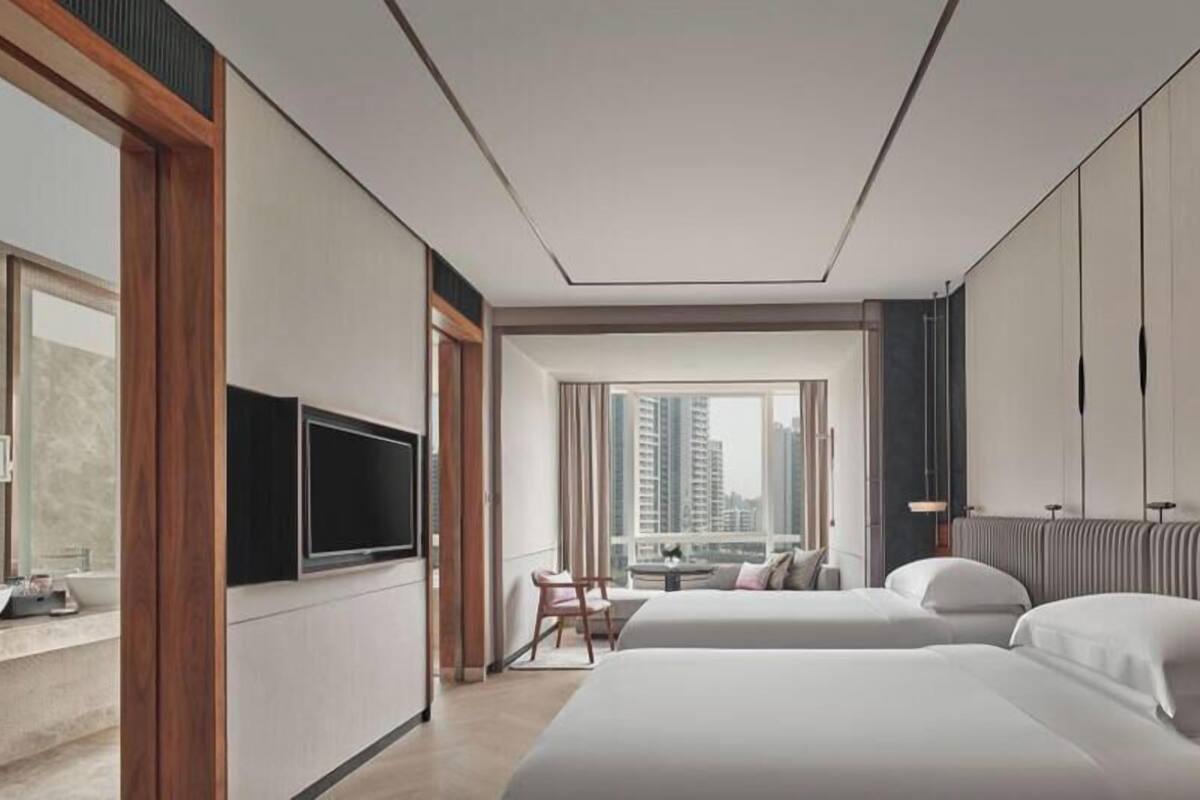 A modern hotel room with two beds, a TV, a view of the city, and a dining area.