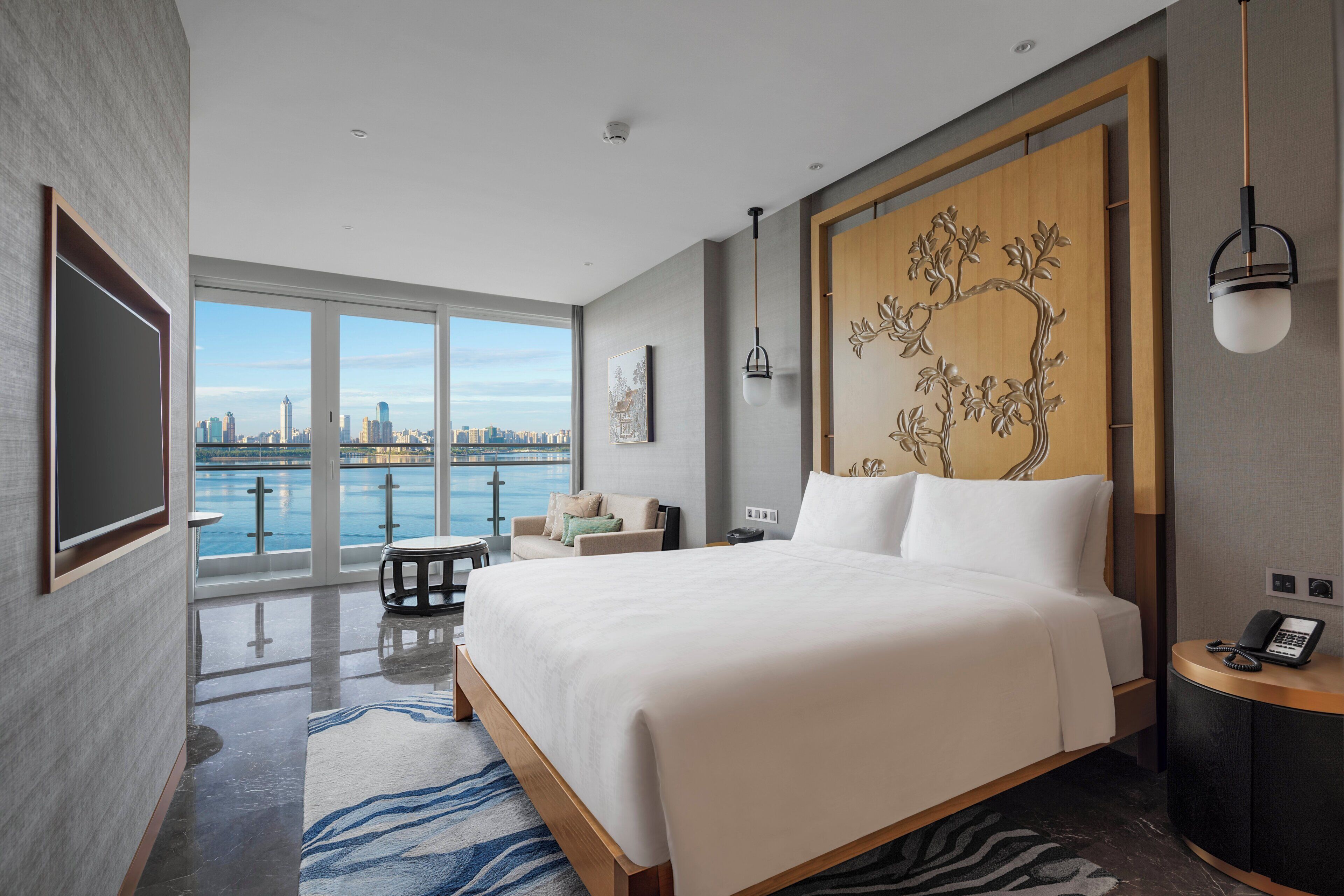 A modern hotel room with a large bed, a view of the cityscape, a balcony with a view of the water, and a wooden accent wall.