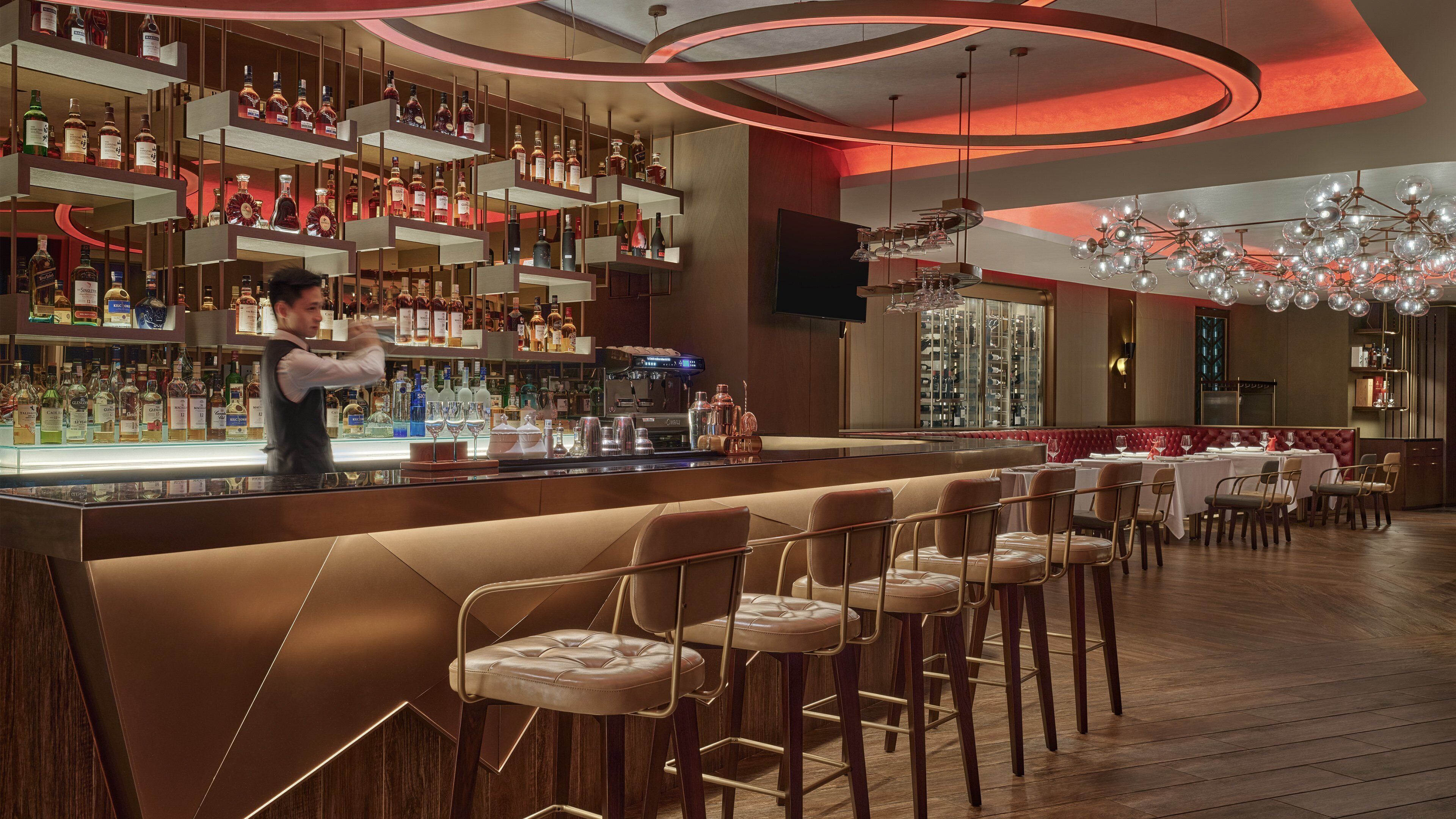 A bar area with a bartender, shelves of liquor bottles, a coffee machine, and a seating area with tables and chairs.