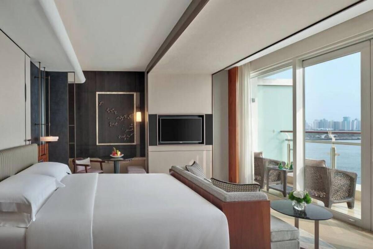 A hotel room with a large bed, a TV, a balcony with a view of the city and water, and a small seating area.