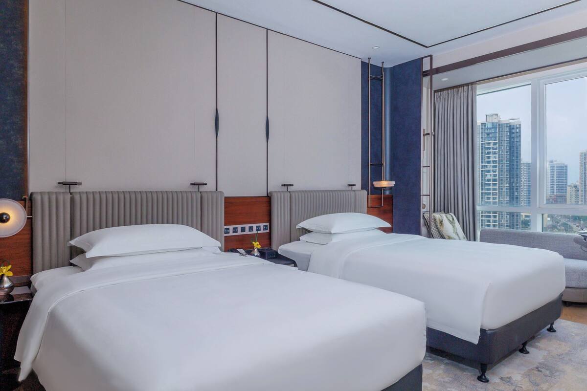 A hotel room with two beds, a large window with a city view, a sofa, and a desk with a lamp.