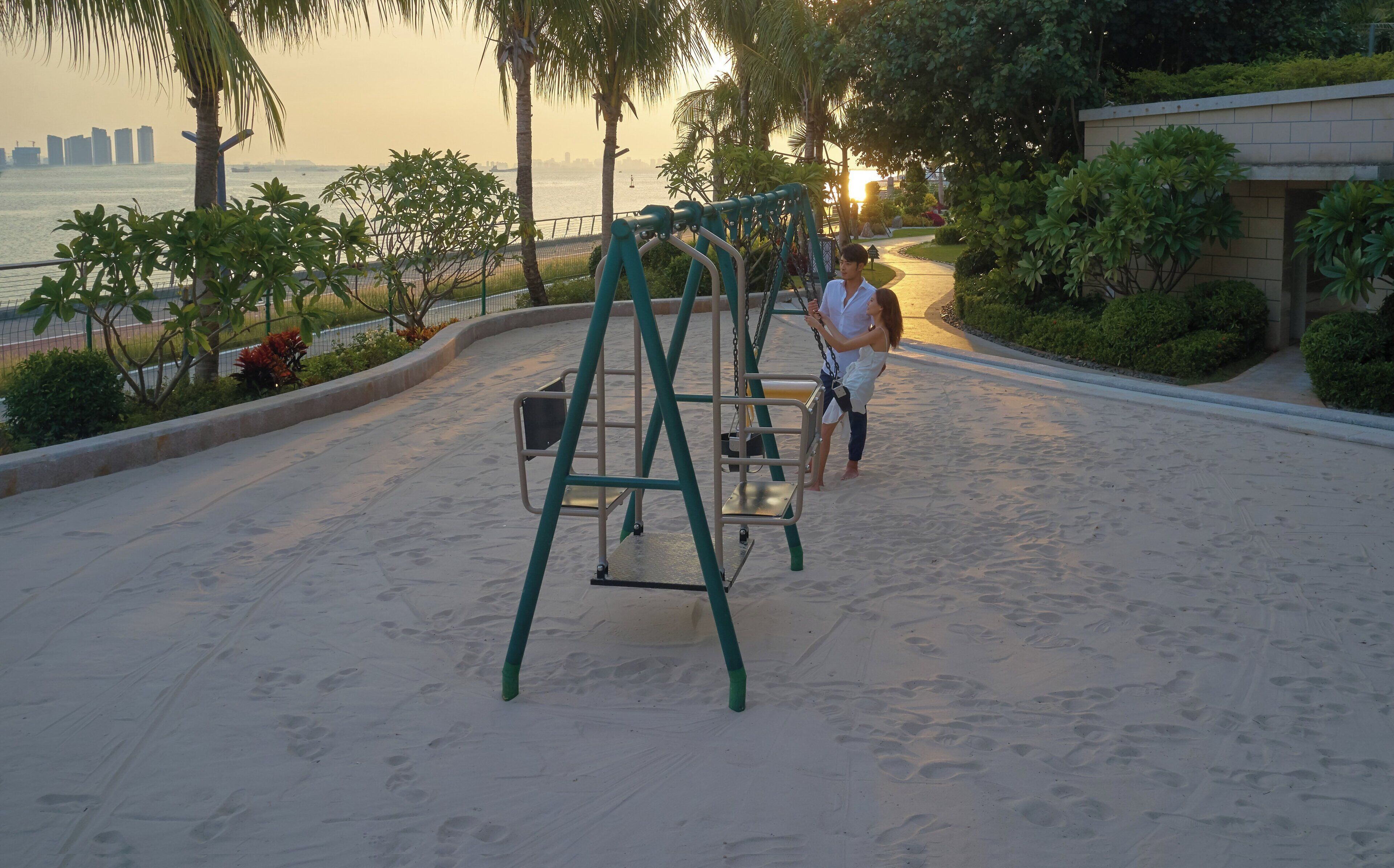 A sandy pathway with exercise equipment and greenery, leading to a body of water with buildings in the distance.