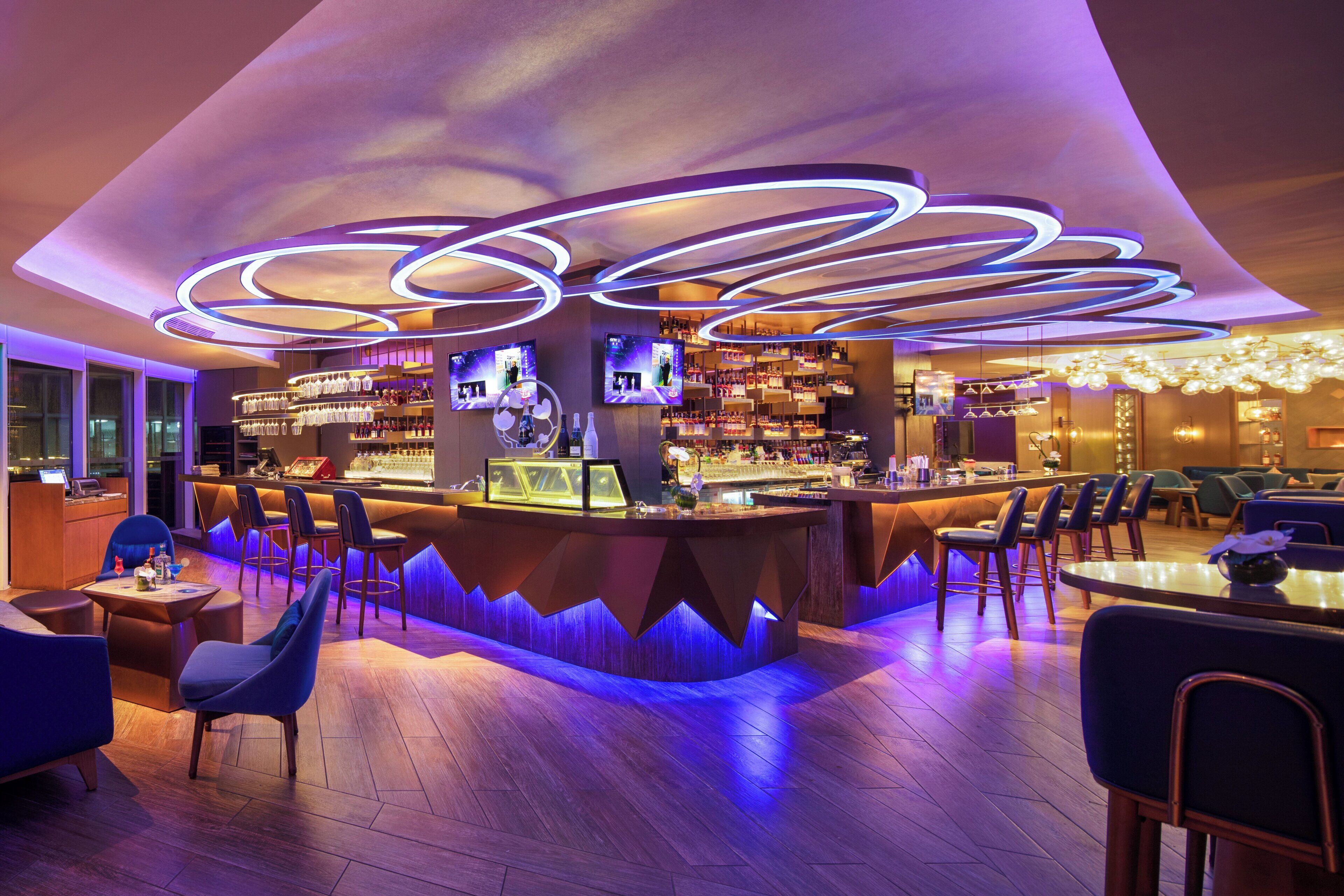 A modern bar area with a curved counter, multiple screens, and a variety of seating options including bar stools and tables.