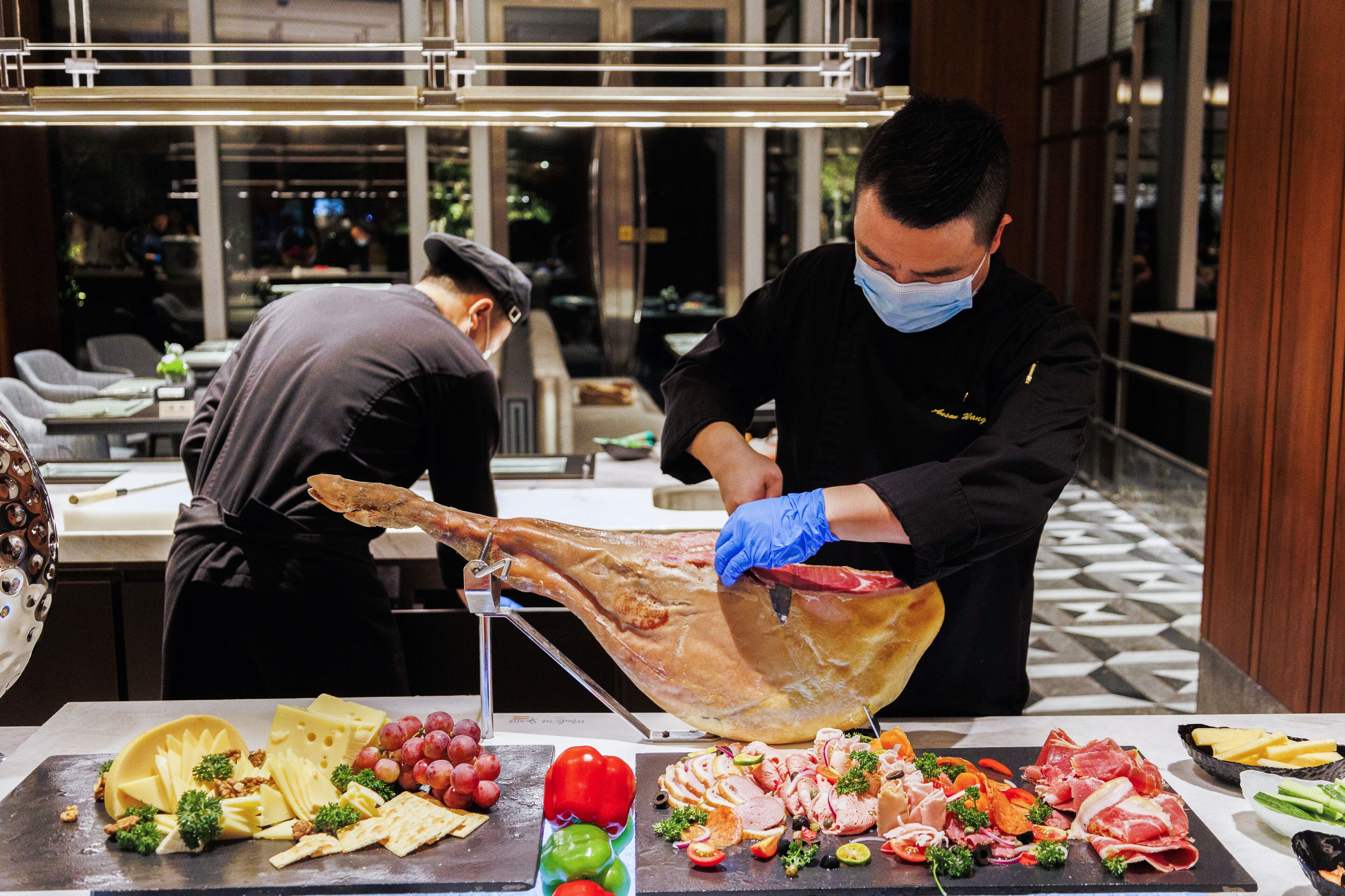 A chef carving a large piece of meat at a buffet table with various cheeses, fruits, and vegetables.