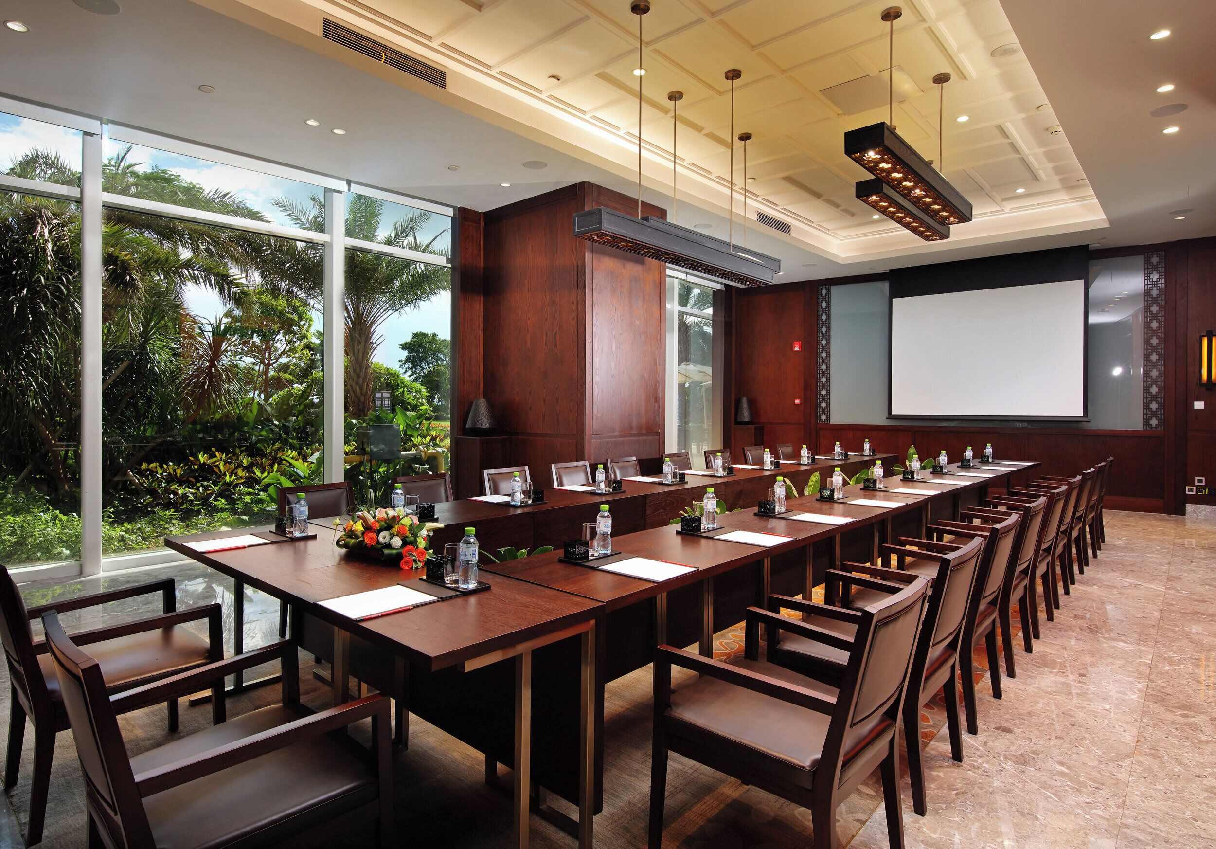 A conference room with a long table, chairs, a projector screen, and a view of greenery outside.