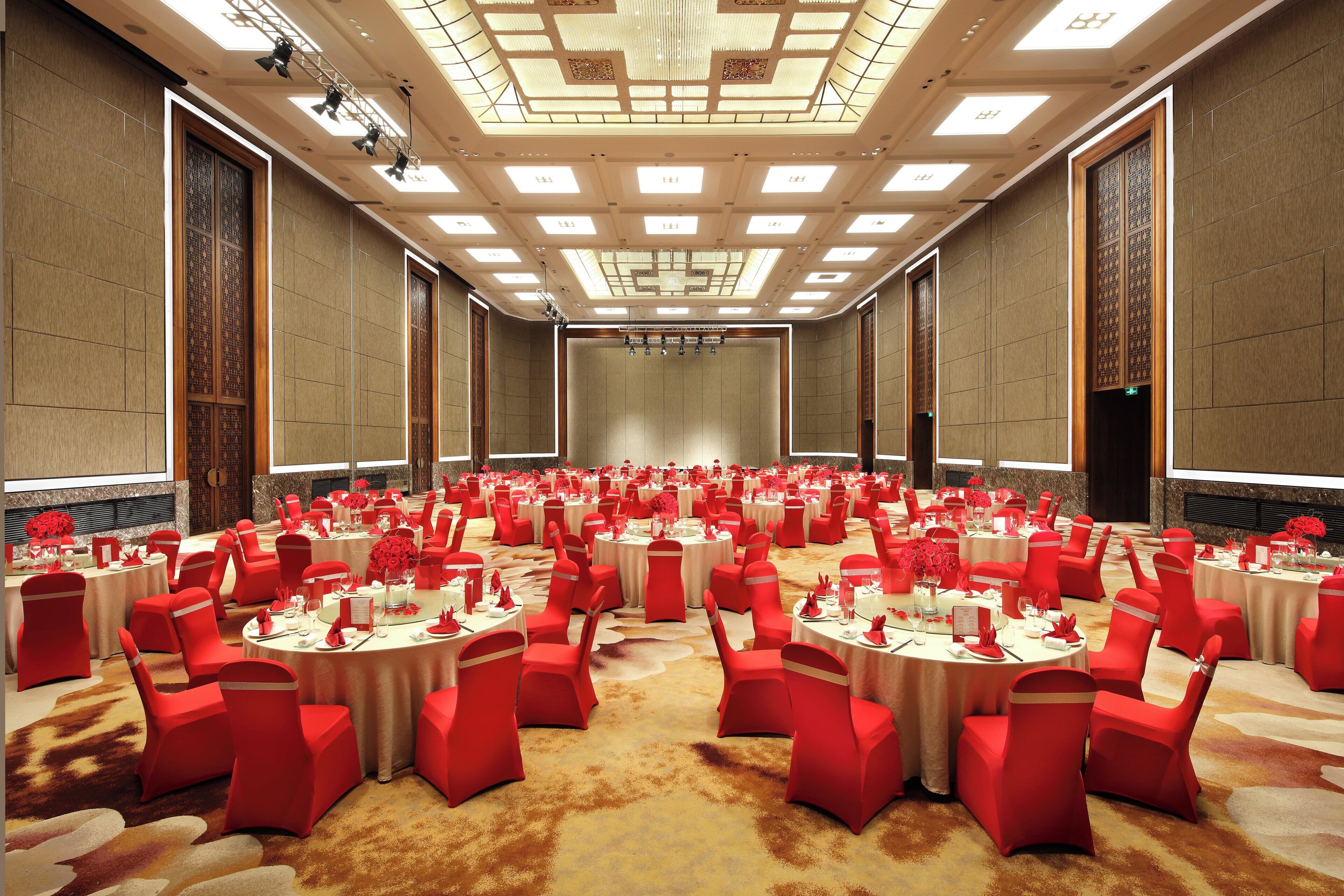 Un amplio salón de conferencias con mesas redondas y sillas rojas, equipado con iluminación empotrada y un diseño de techo estampado.