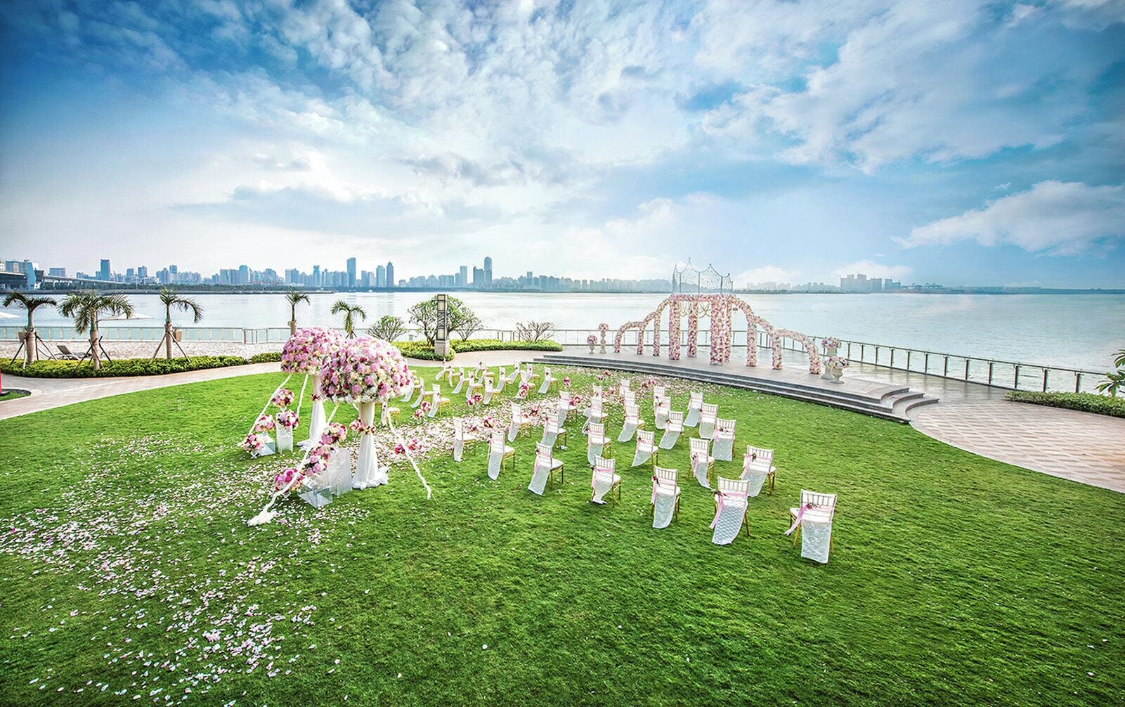 A wedding ceremony setup with chairs arranged in a semi-circle, a floral arch, and a view of a city skyline across a body of water.