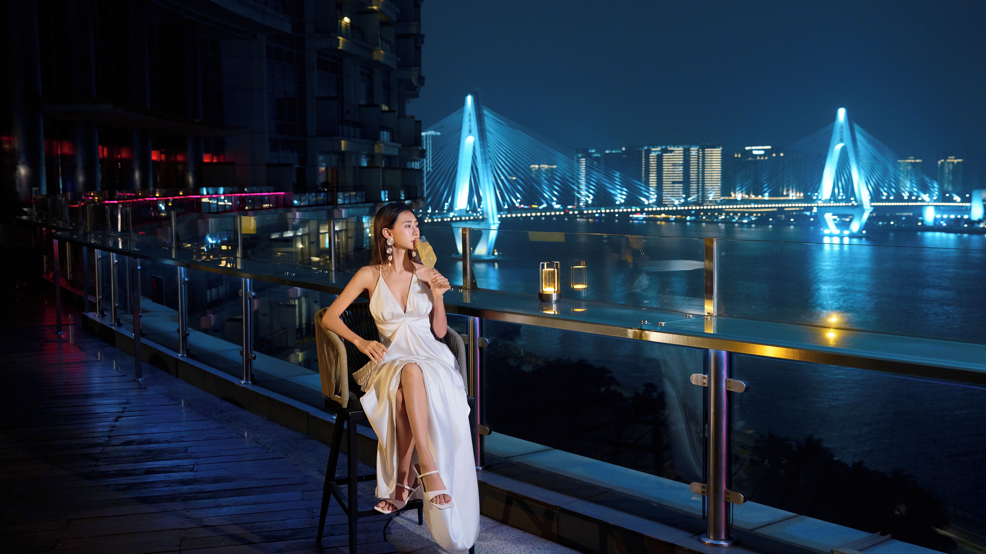A woman seated on a chair by a waterfront with a bridge in the background.