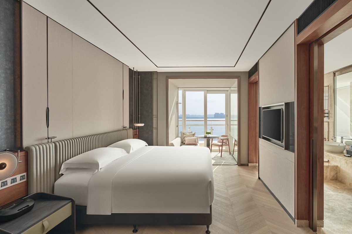 A modern hotel room with a large bed, a TV, a desk, and a view of the water.