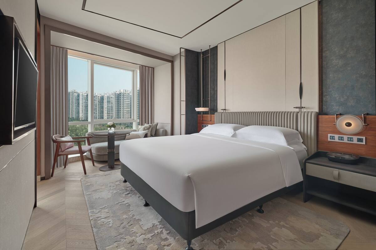 A modern hotel room with a large bed, a desk, and a view of high-rise buildings.