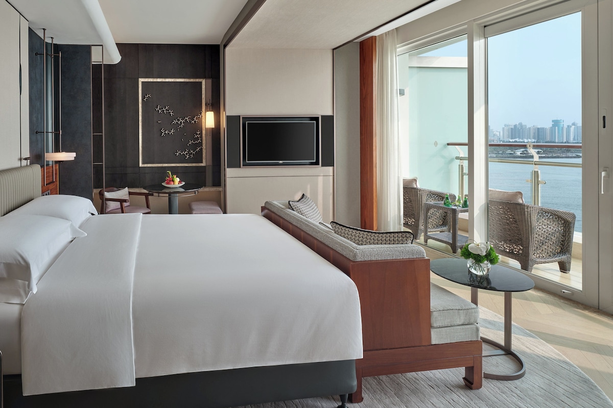 Una habitación de hotel moderna con una cama grande, una mesita con frutero, un sofá y un balcón con vistas a la ciudad.