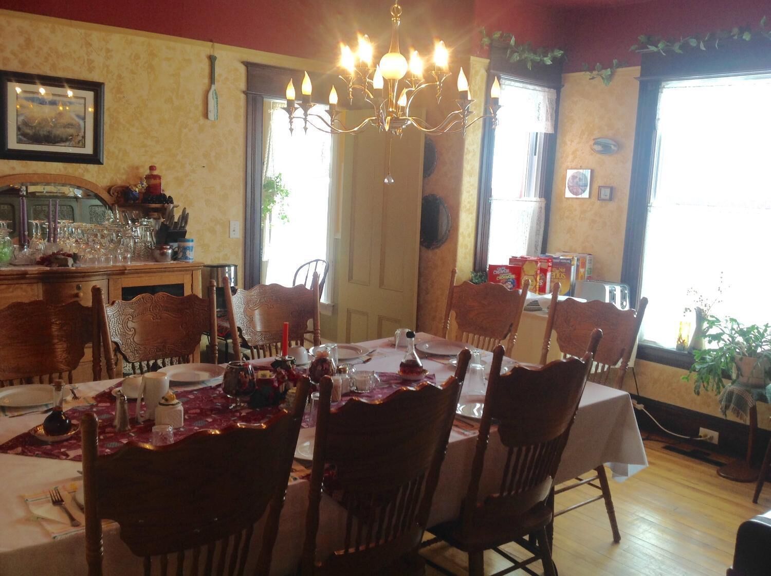 A dining area with a chandelier, wooden chairs, a table set for a meal, and a window with plants.