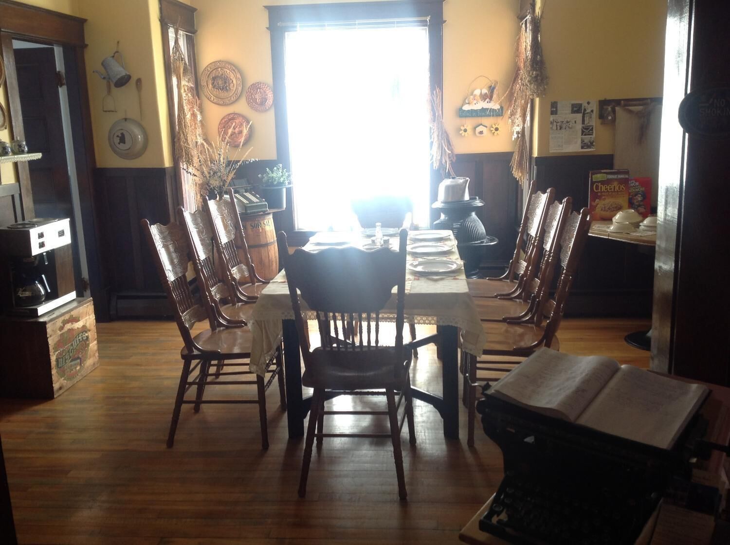A dining area with a wooden table set for four, surrounded by wooden chairs. A coffee machine is on the left, and a box labeled "Cheerios" is on the right.