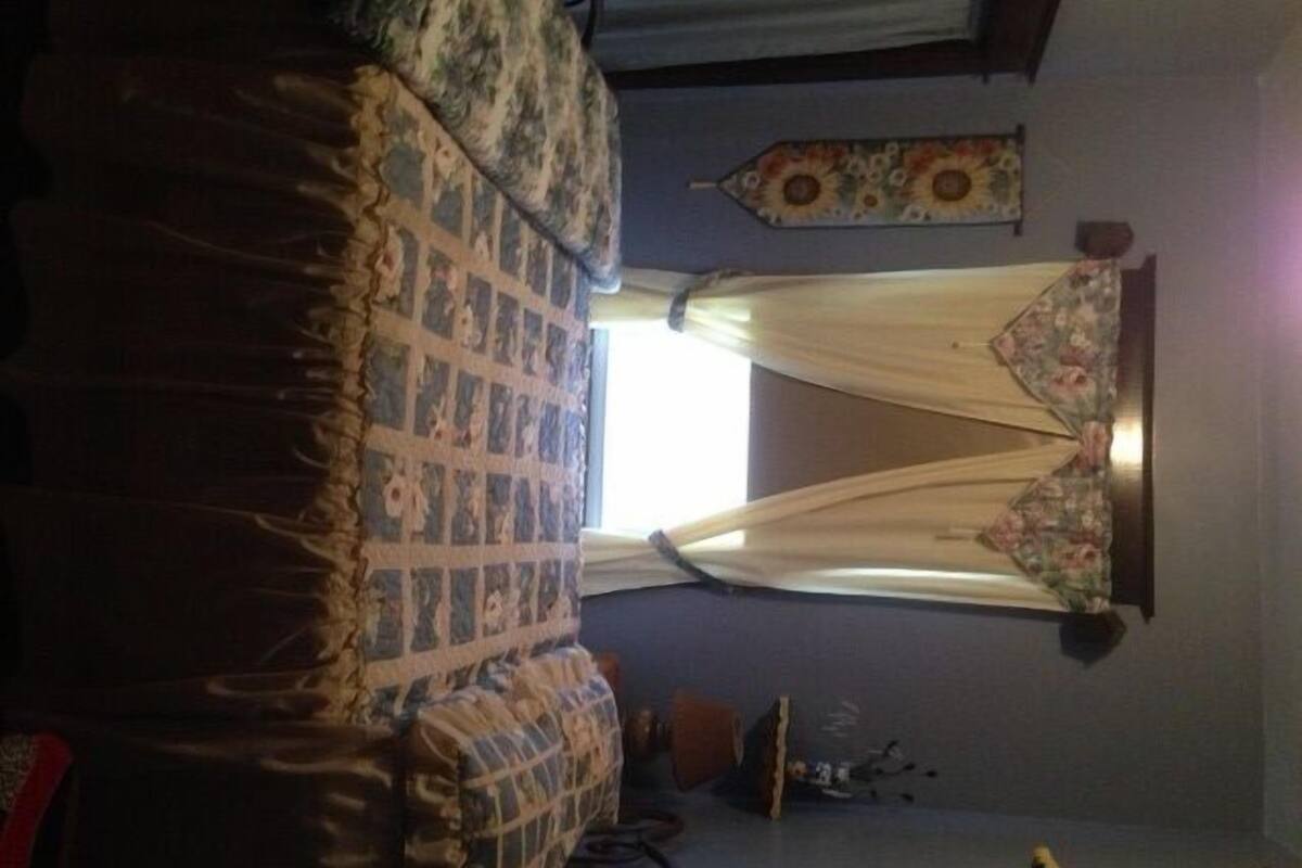 Une chambre à coucher avec un lit, une fenêtre avec des rideaux, un élément de décoration murale et une petite table sur laquelle sont posés divers objets.