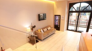 A modern hotel lobby with a beige sofa, a mounted TV, a vending machine, and a staircase.