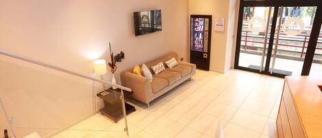 A modern hotel lobby with a beige sofa, a mounted TV, a vending machine, and a staircase.