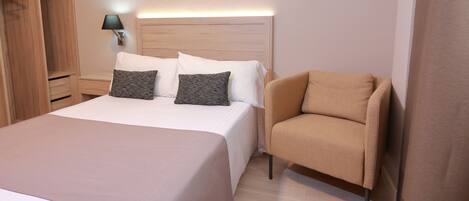 Una habitación de hotel con una cama grande, un armario de madera, una mesita de noche con lámpara y un sillón beige.