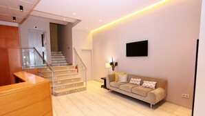 A modern living room with a wooden reception desk, a beige sofa with patterned cushions, a mounted flat-screen TV, and a staircase with glass railings leading to an upper floor.