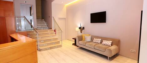 A modern living room with a wooden reception desk, a beige sofa with patterned cushions, a mounted flat-screen TV, and a staircase with glass railings leading to an upper floor.