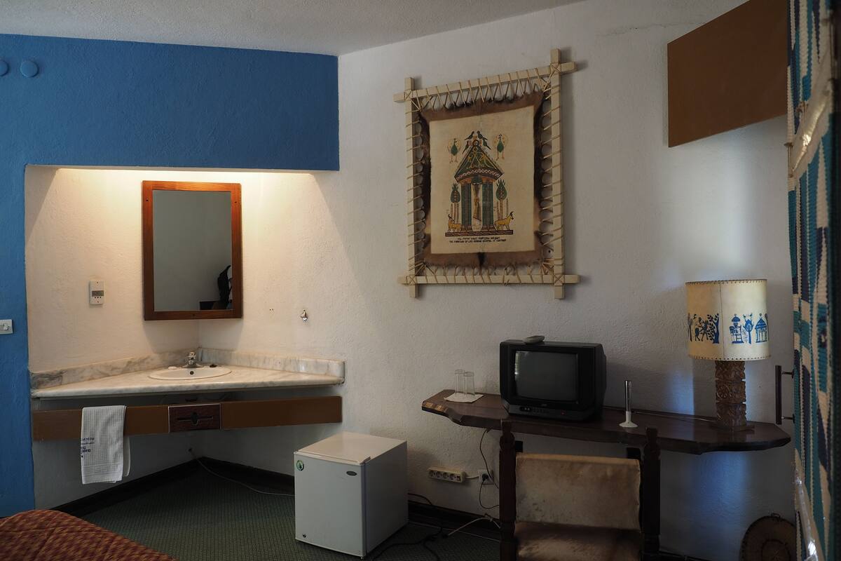 A compact hotel room with a small sink, a mirror, a television, a wooden table, and a decorative wall hanging.