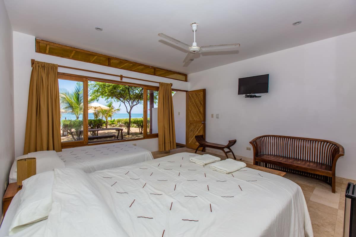 Habitación de hotel con dos camas, ventilador de techo, televisión y vista a la playa.