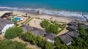 Vista aérea de un resort en la playa con cabañas de techo de paja, una piscina y una vista despejada del océano.