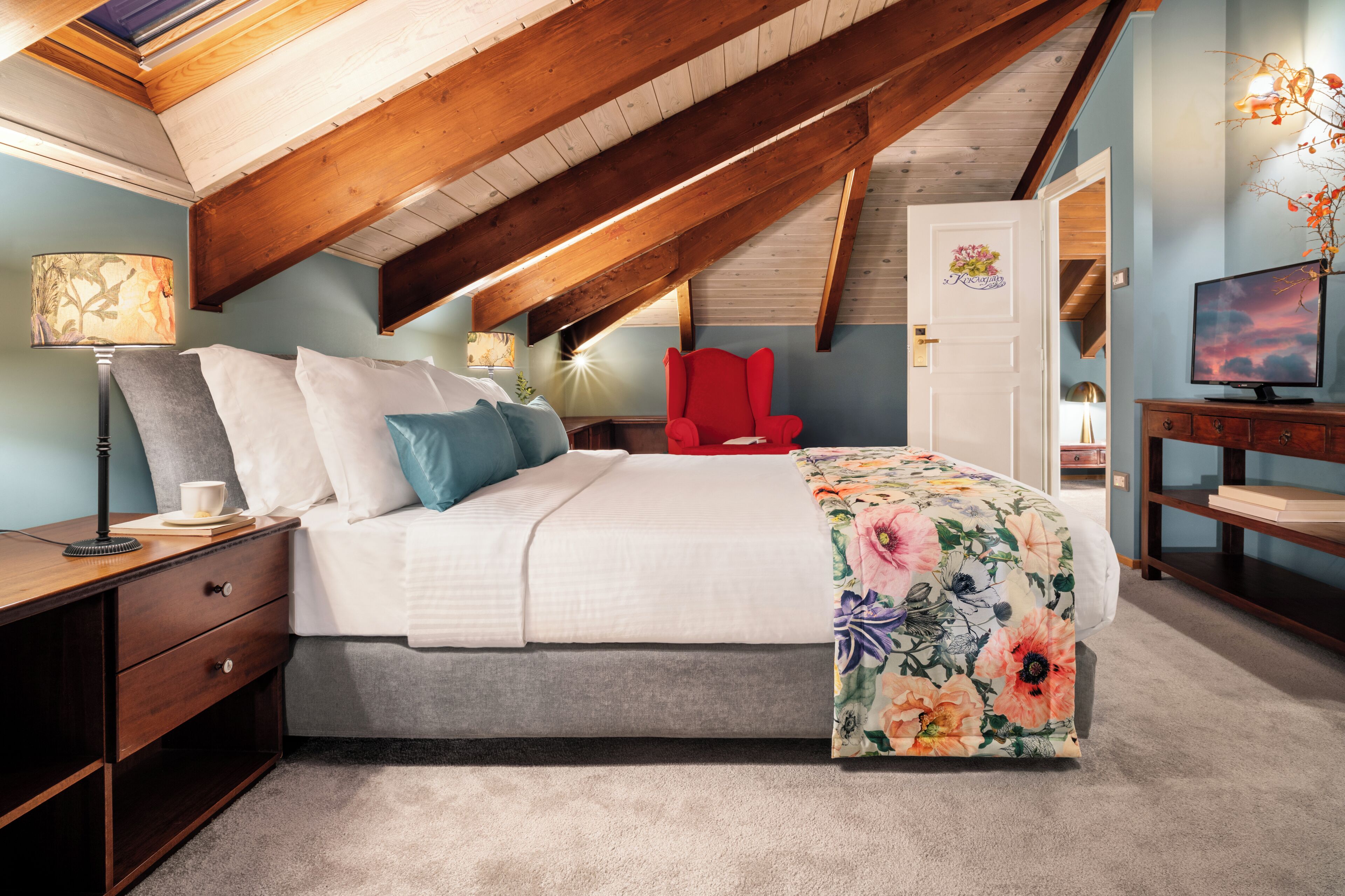 A bedroom with a sloped ceiling, a bed with a floral bedspread, a red chair, a TV, and a desk with a lamp.