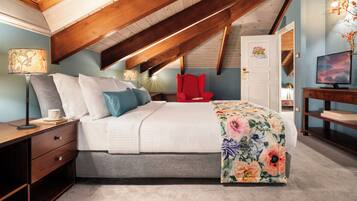 A bedroom with a sloped ceiling, a bed with a floral bedspread, a red chair, a TV, and a desk with a lamp.
