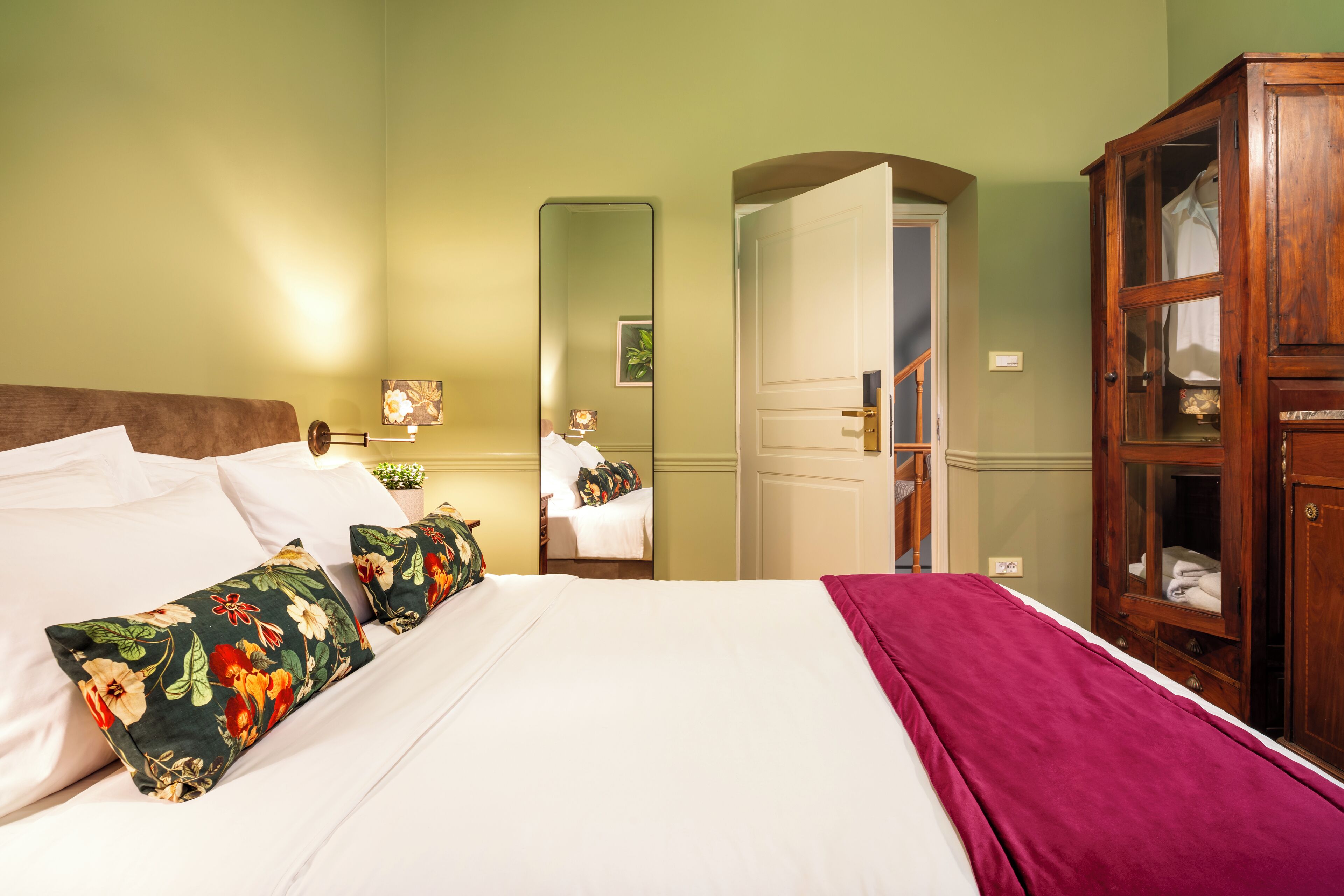 A neatly made bed with floral pillows, a wooden wardrobe, and a mirror reflecting a room with a door and a plant.